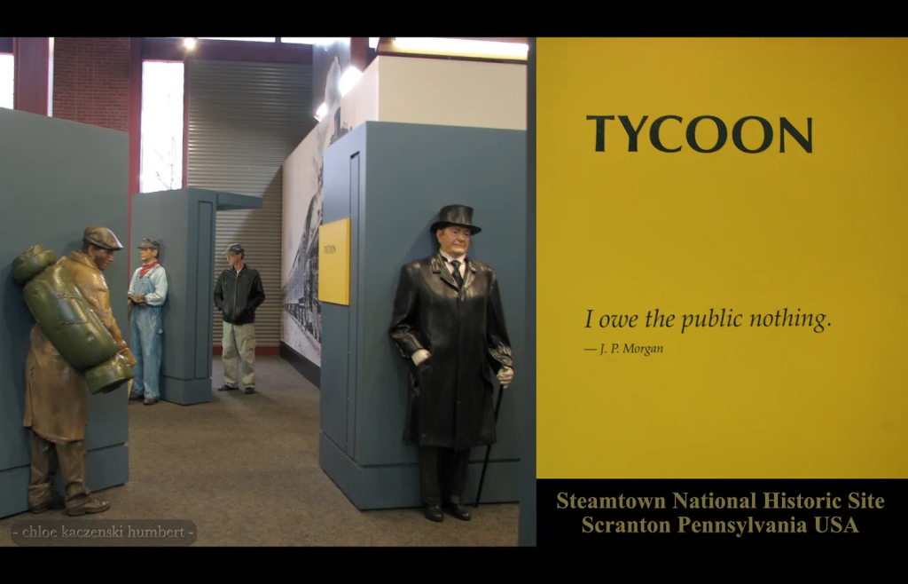 The image is 2 photos, one is of a man in a baseball cap, black leather jacket and tan cargo pants standing in a museum looking at a display among life size statues of a shabbily dressed man with a duffle bag, trench coat and golf cap and with a demeanor of tiredness, another statue of a man in light blue overalls and a red scarf and a train conductor hat standing straight back against a wall, and the third statue is of a businessman leaning back against a wall all in a black top hat, black long coat, black pants, black boots, a white shirt with a black tie, wearing white gloves and holding a black cane, and looking downward with an expression of boredom. Next to the top hat man is a yellow sign that is at an angle where it can’t be read, so the image has a closeup next to the photo showing the yellow sign which has a one line quote from J.P. Morgan saying I owe the public nothing. The caption on the image says Steamtown National Historic Site Scranton Pennsylvania USA. photo by chloe kaczenski humbert